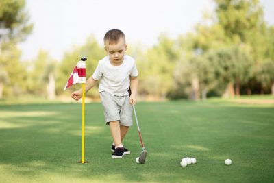 little Boy playing golf and hitting ball by putter on green grass.